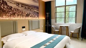A hotel room with a large bed, a cityscape mural, a small table, two chairs, and a window with a view of greenery. - Dinghu Tiantian Business Hotel (Zhaoqing)