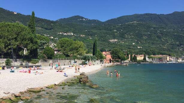 A beach with clear water, rocky shore, and buildings in the background. - Casa Fornico 2, with terrace and partial lake view in quiet position (Lombardia)