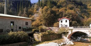A stone building with a red roof, a white building with a red roof across a bridge, and a river with rocks. - Casa Fornico 2, with terrace and partial lake view in quiet position (Lombardia)