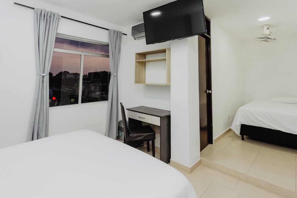 A hotel room with a bed, a desk with a chair, a TV mounted on the wall, and a window with curtains.
