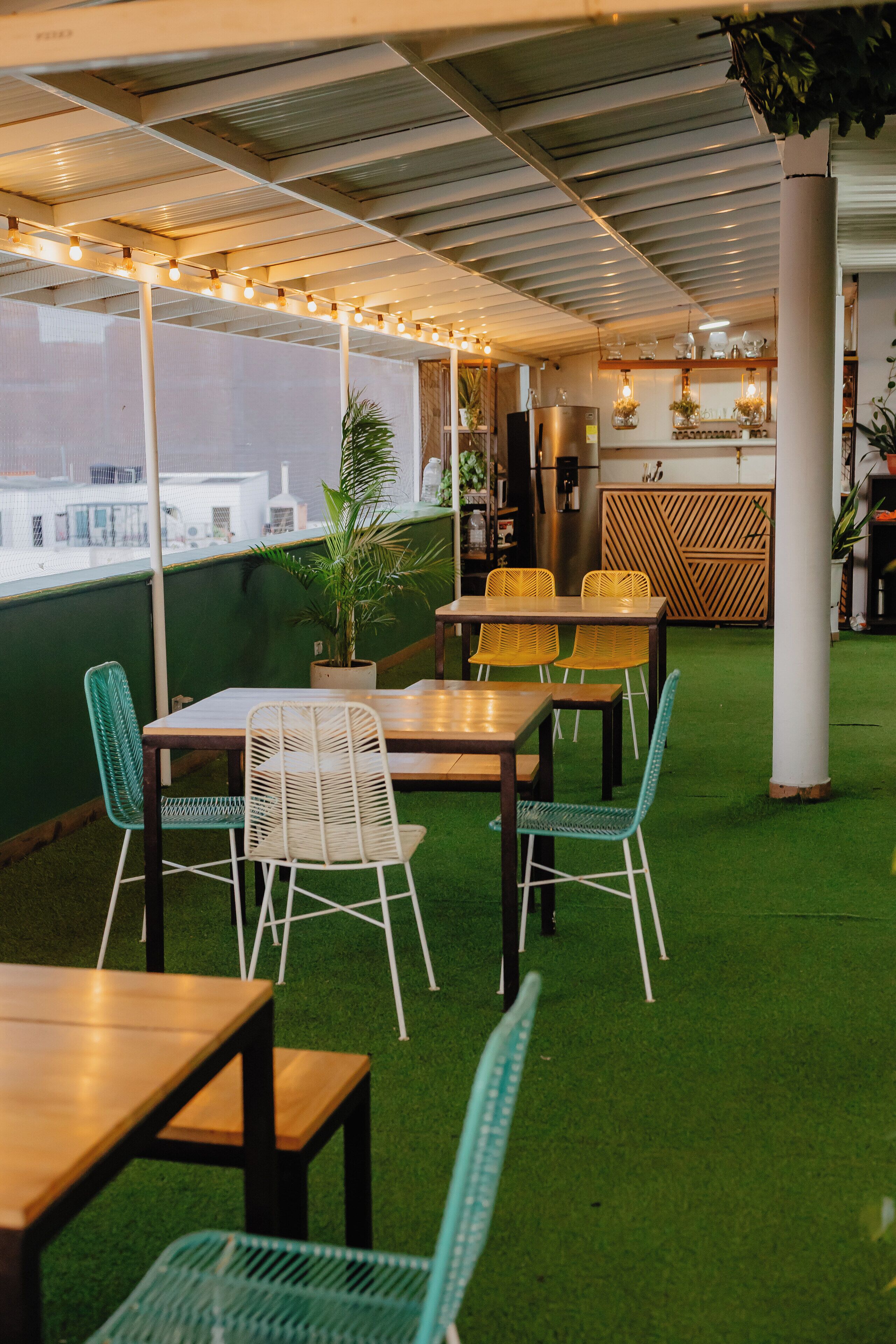 An indoor seating area with tables and chairs, a green artificial turf floor, and a view of buildings through a window.