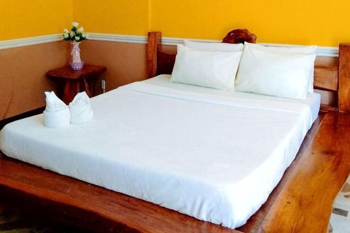 A neatly made bed with white linens and pillows, a wooden headboard, and a small wooden table with a vase of flowers beside it.