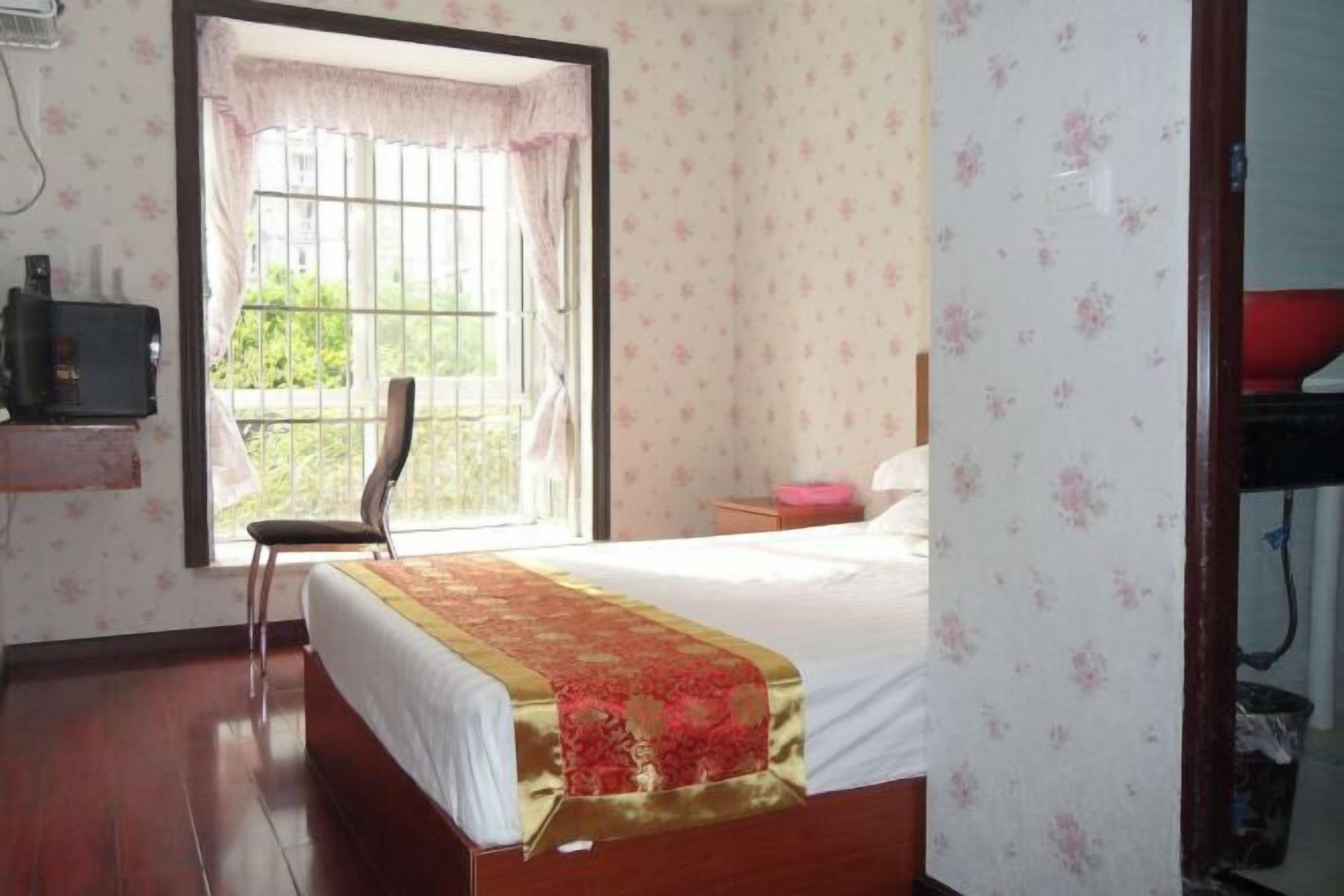 A bedroom with a bed, a chair, a window with curtains, a bedside table, and a wall-mounted TV.