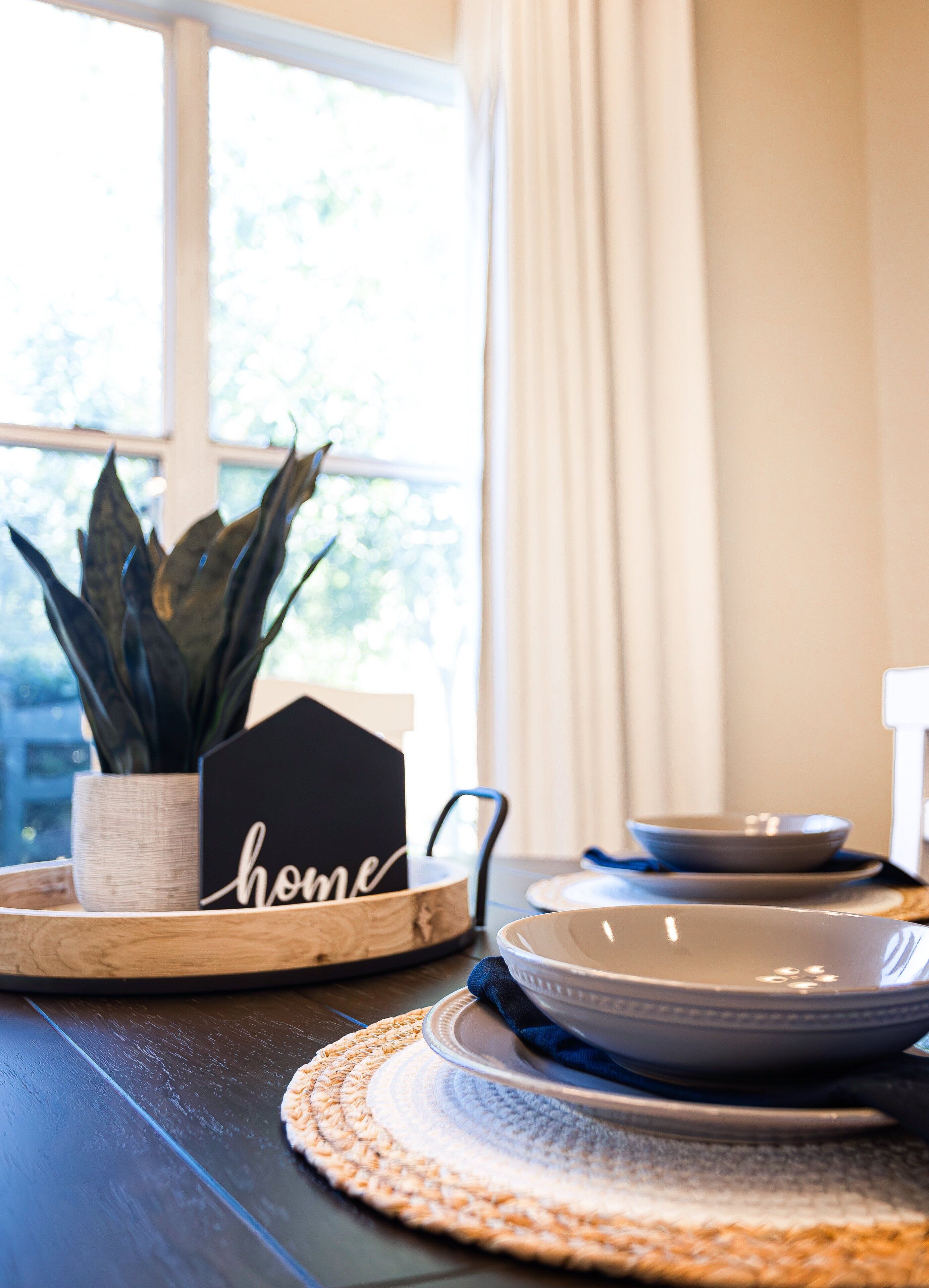 Una mesa de comedor con un posavasos, una planta en maceta, un cartel con la palabra 'home' y cuencos y platos apilados.