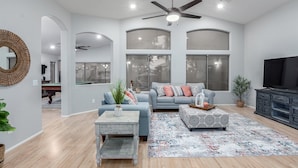 A spacious living room with a ceiling fan, large windows, a sofa, a coffee table, and a TV. - 165th Goodyear (Goodyear)