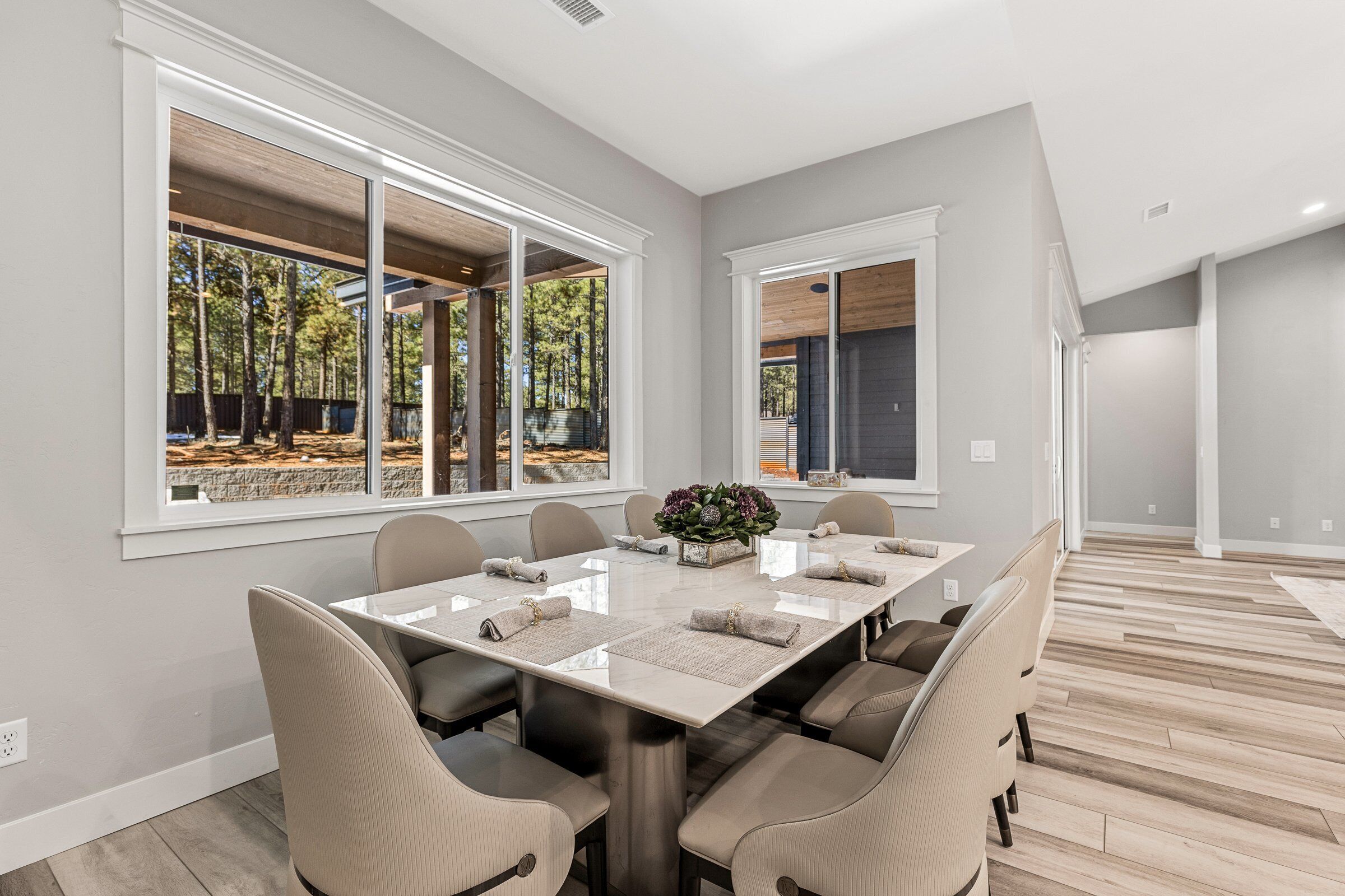 A modern dining room with a large table, comfortable chairs, and large windows offering a view of trees.