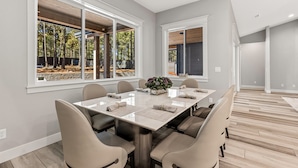 A modern dining room with a large table, comfortable chairs, and large windows offering a view of trees.