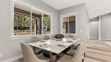A modern dining room with a large table, comfortable chairs, and large windows offering a view of trees.