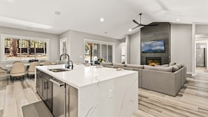 A modern kitchen with a large island, a dining area with chairs, and a living room with a sofa and fireplace.