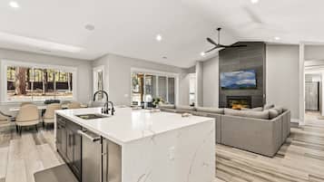 A modern kitchen with a large island, a dining area with chairs, and a living room with a sofa and fireplace.