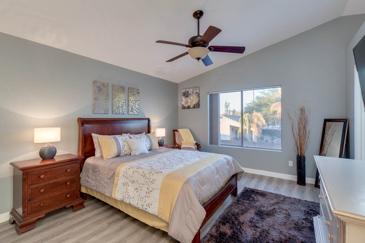 A bedroom with a bed, nightstands, a ceiling fan, a window, and artwork on the walls.