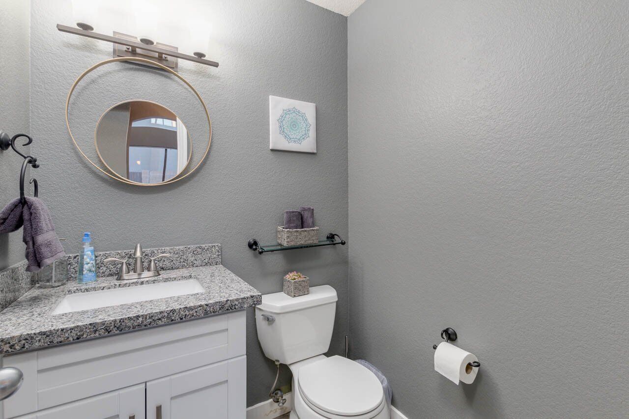 A bathroom with a granite countertop sink, a round mirror, a toilet, and a towel hanging on a hook.