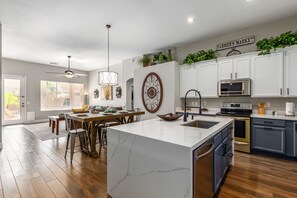 A modern kitchen with white cabinets, a large island, and a dining area with wooden chairs and a table. - Harrison Chandler (Chandler)