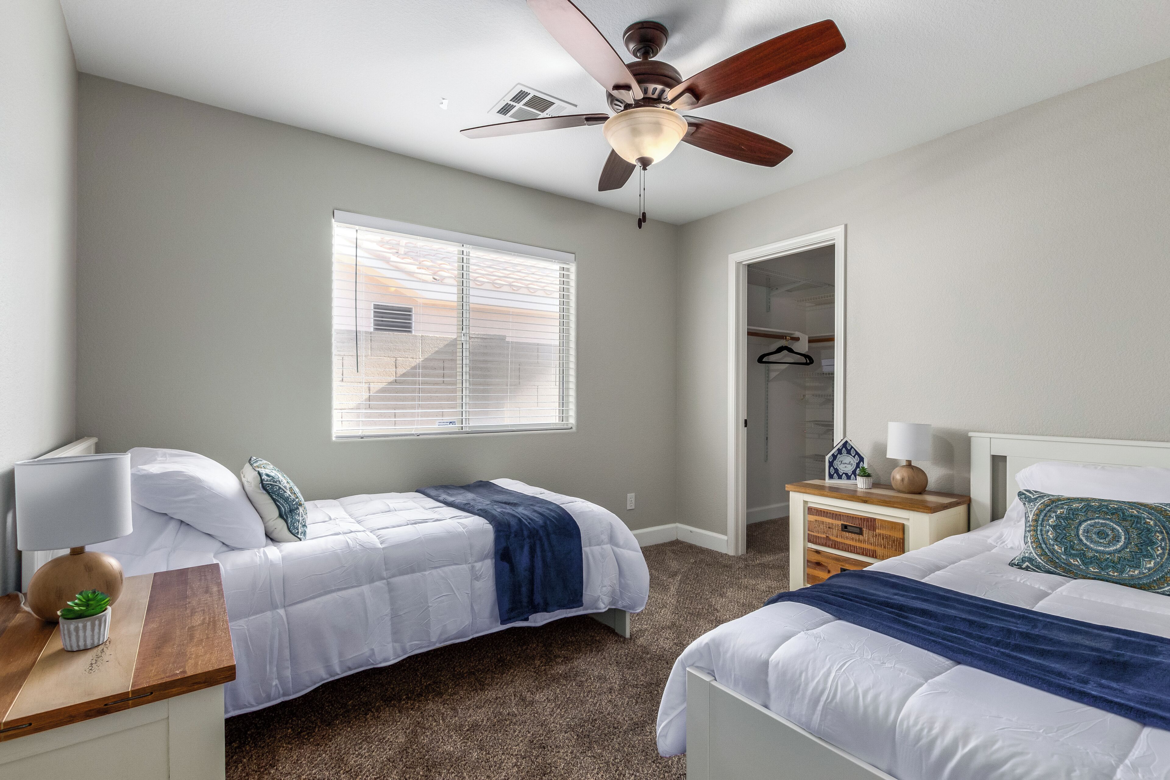 A bedroom with two beds, a ceiling fan, a window with blinds, a nightstand, and a walk-in closet.