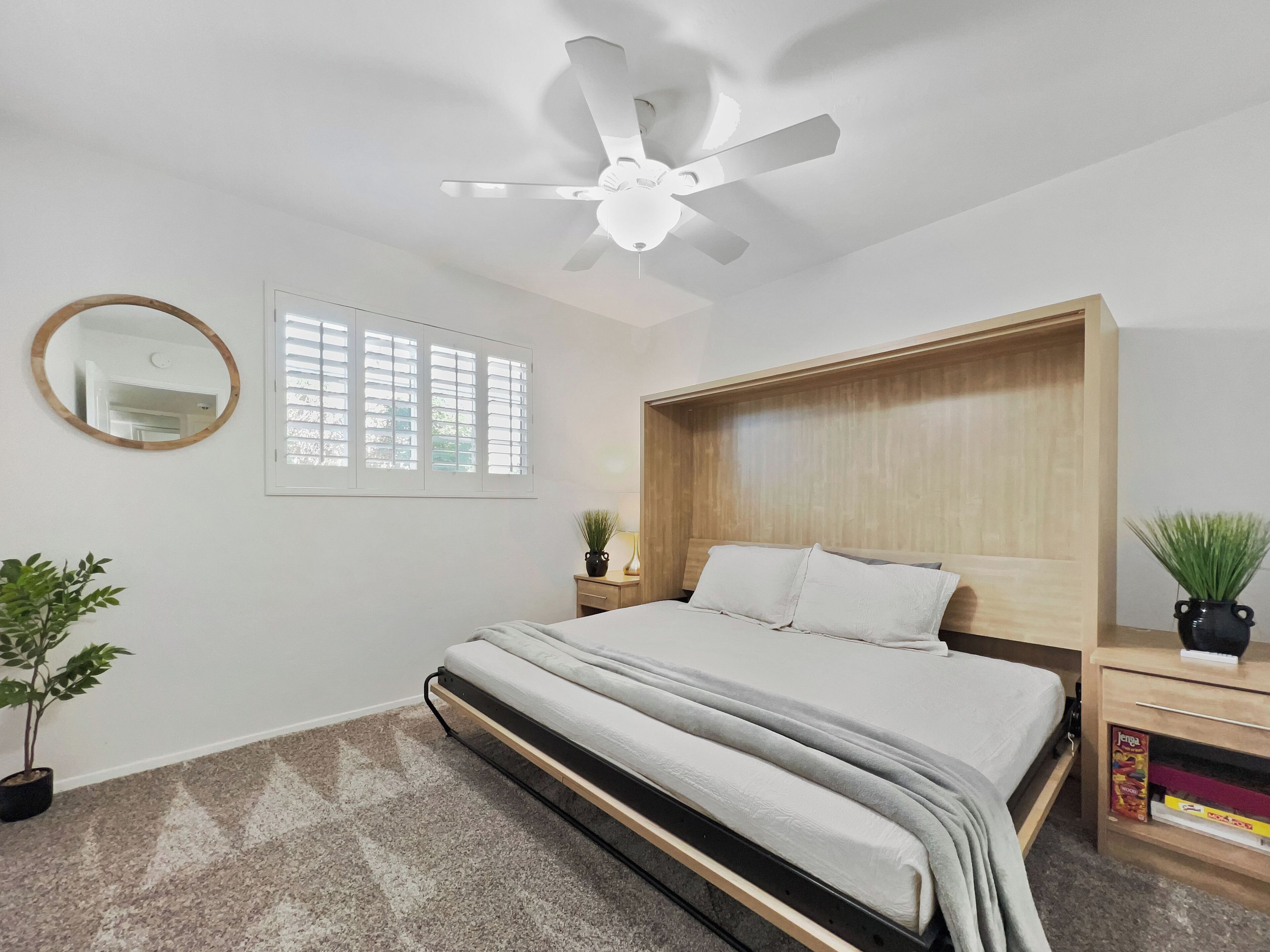 A bedroom with a bed, a ceiling fan, a window with blinds, a round mirror, and a bedside table with a plant.