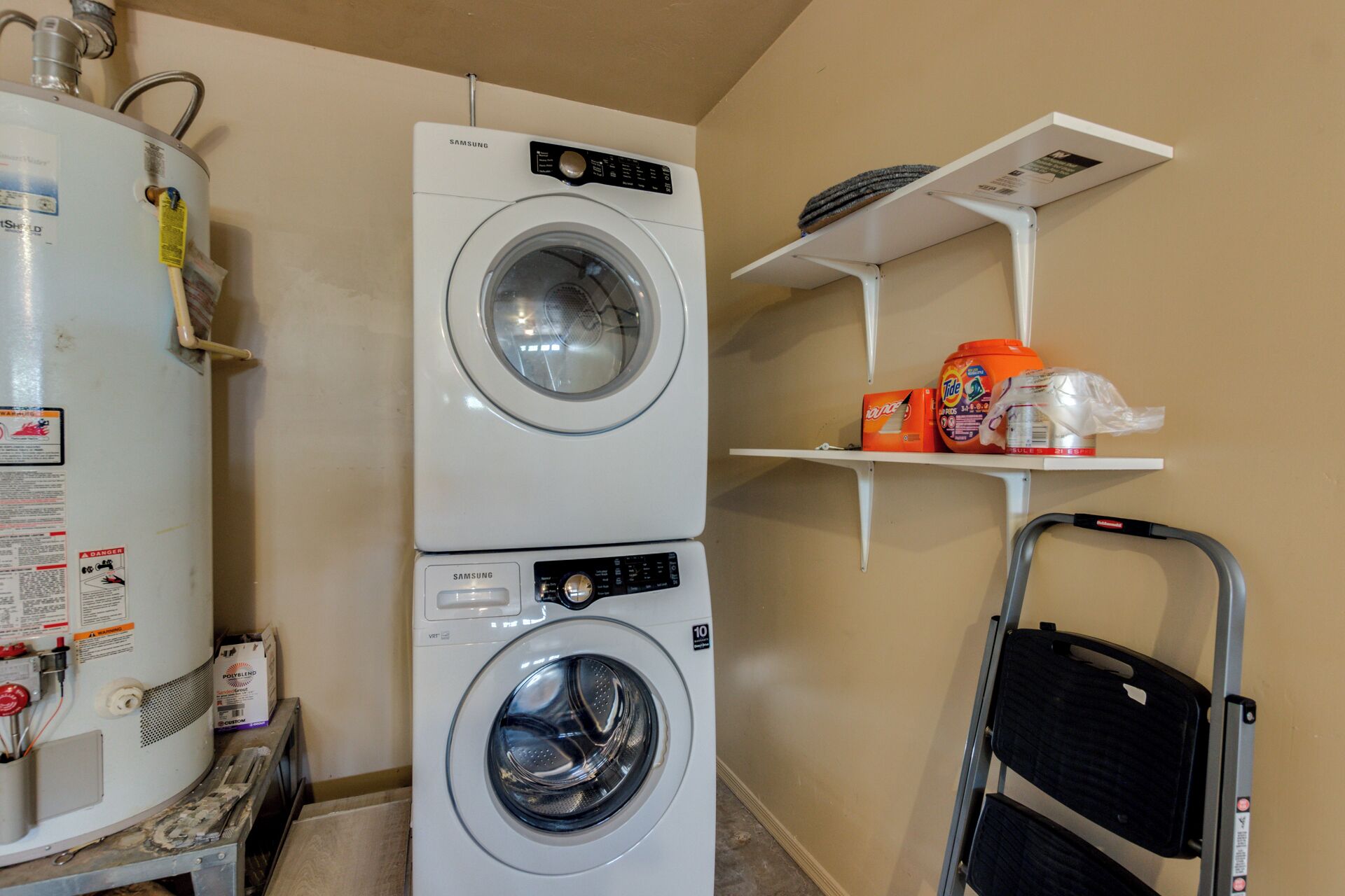 A laundry area with two stacked washers and dryers, a water heater, and shelves with laundry supplies.