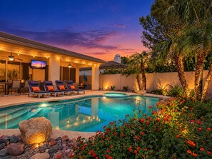 A swimming pool with lounge chairs and a television screen, surrounded by a landscaped garden with rocks and flowering plants. - Lynx Chandler/ocotillo (Chandler)