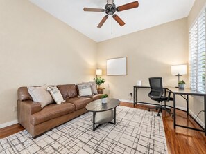 Un salon avec un canapé en cuir marron, une table basse, un ventilateur de plafond, un bureau avec une chaise et une fenêtre avec des stores.