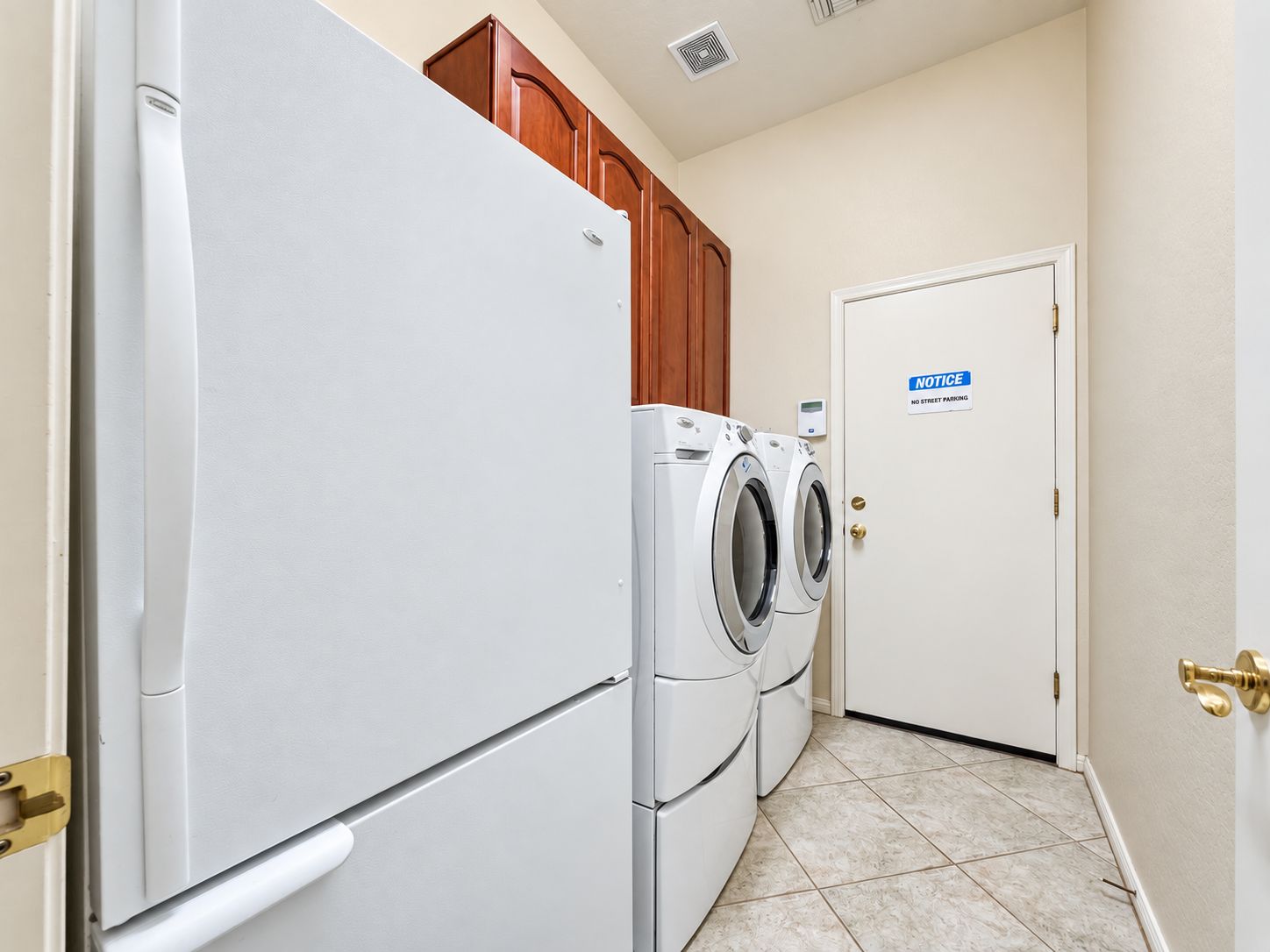A laundry room with a white refrigerator, a washing machine, and a dryer. There is a door with a 'No Street Parking' sign.