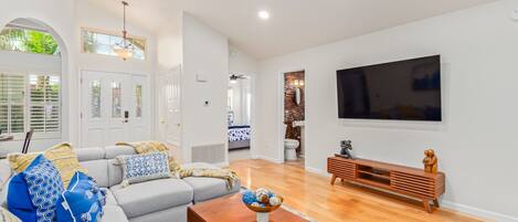 A spacious living room with a grey sofa, wooden coffee table, and a TV on the wall.