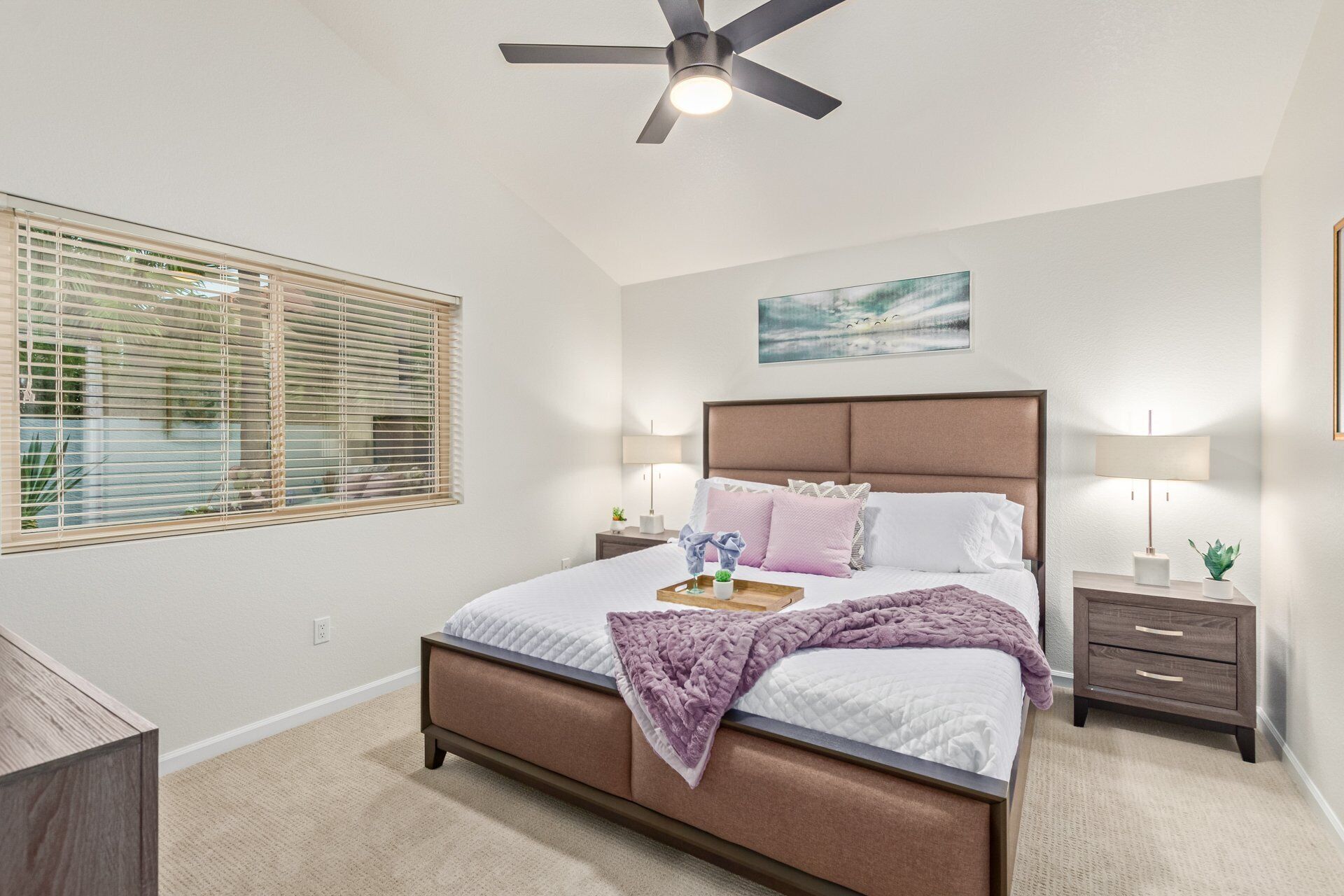 A bedroom with a bed, bedside tables, a ceiling fan, a window with blinds, and a painting on the wall.