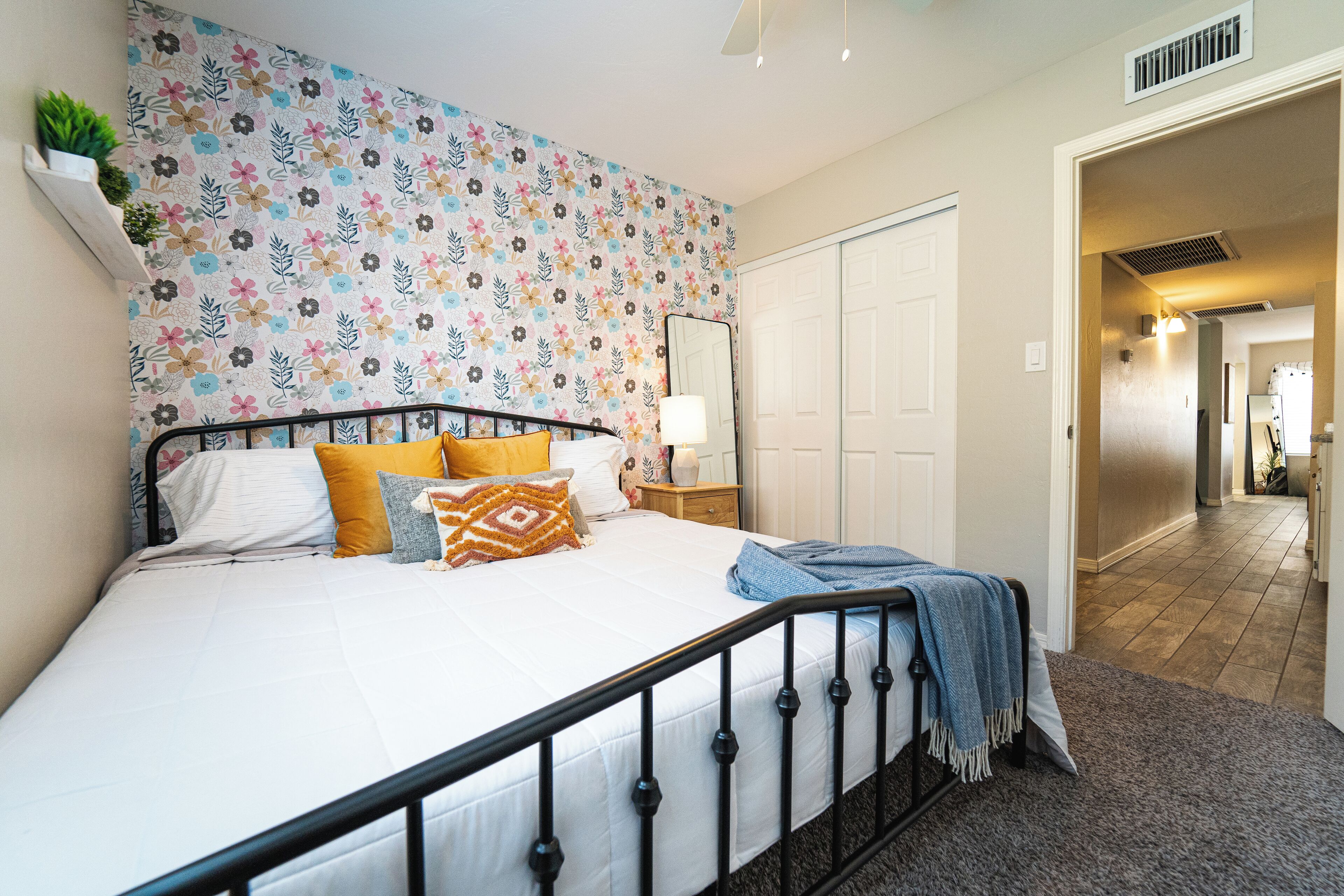 Une chambre avec un mur orné d’un motif floral, un lit recouvert de plusieurs coussins, une table de chevet avec une lampe, et une porte donnant sur une autre pièce.