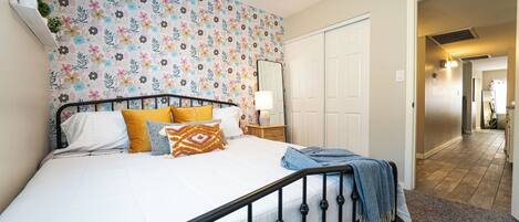 Une chambre avec un mur orné d’un motif floral, un lit recouvert de plusieurs coussins, une table de chevet avec une lampe, et une porte donnant sur une autre pièce.