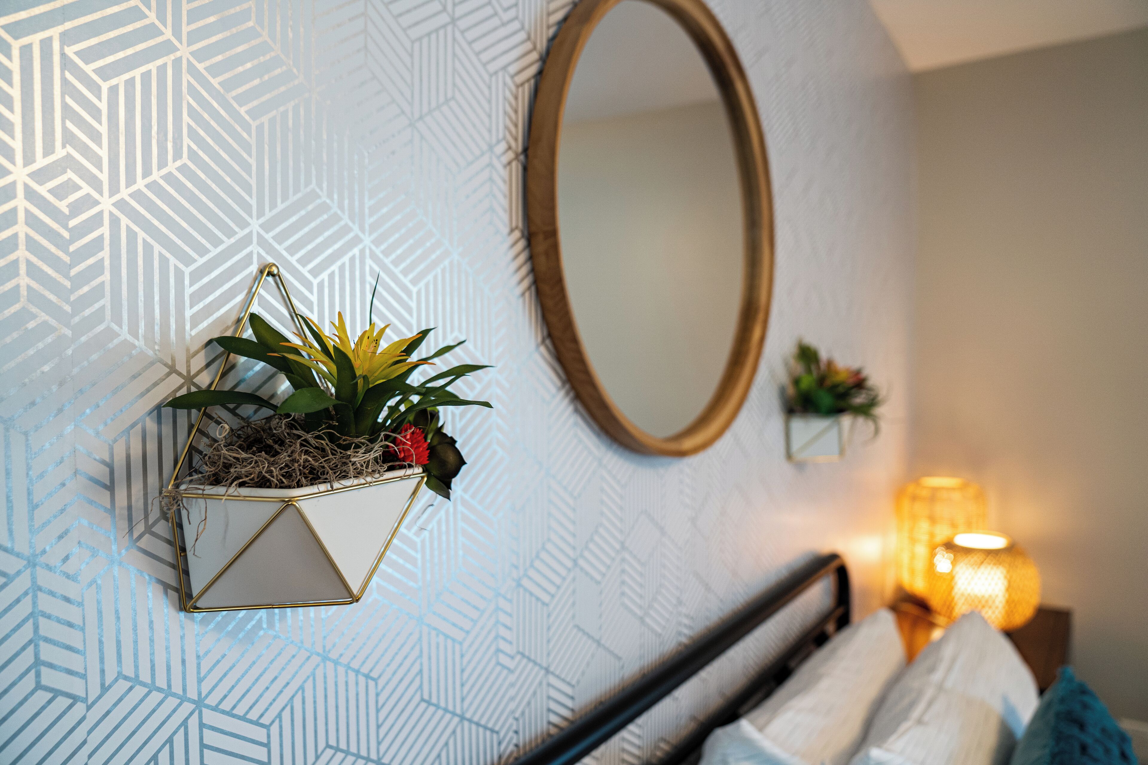 A room with a geometric patterned wallpaper, a wall-mounted mirror, a bedside plant, and a bedside lamp.