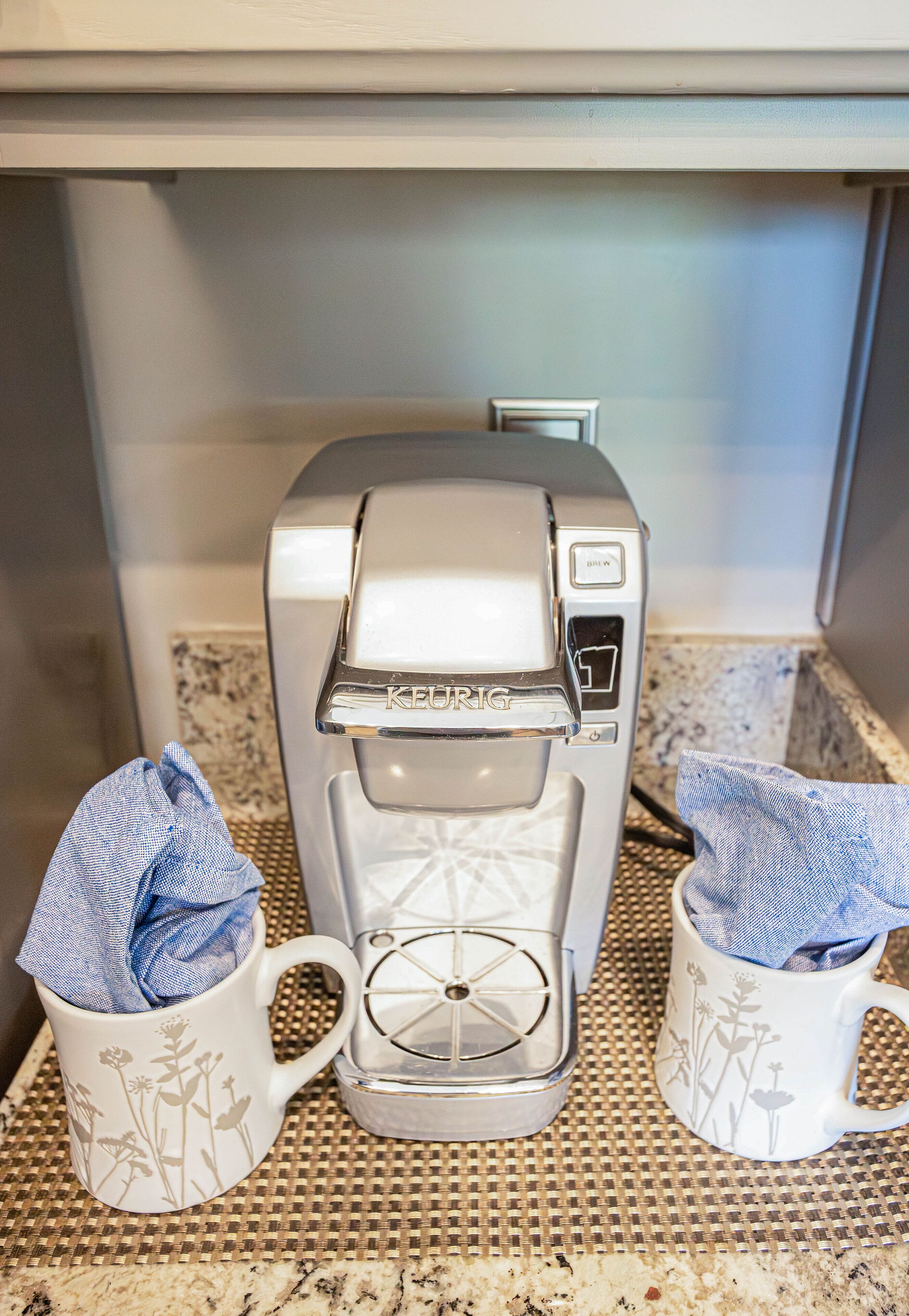 Een Keurig koffiezetapparaat op een keukenblad, met twee koffiemokken, waarvan er één een blauwe handdoekje in heeft.