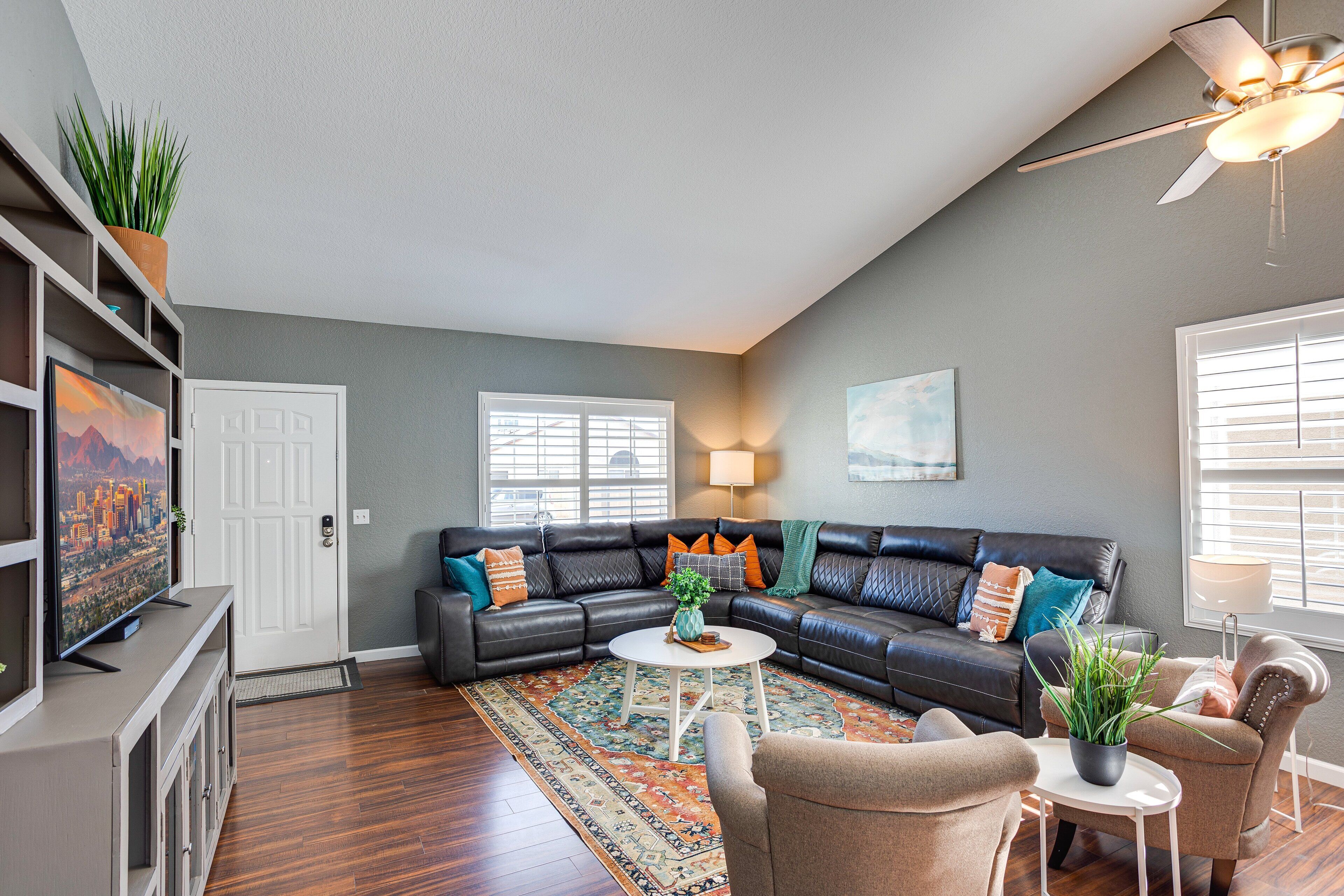 A living room with a sectional sofa, a coffee table, and a TV. There is a ceiling fan, a door, and a window with blinds.