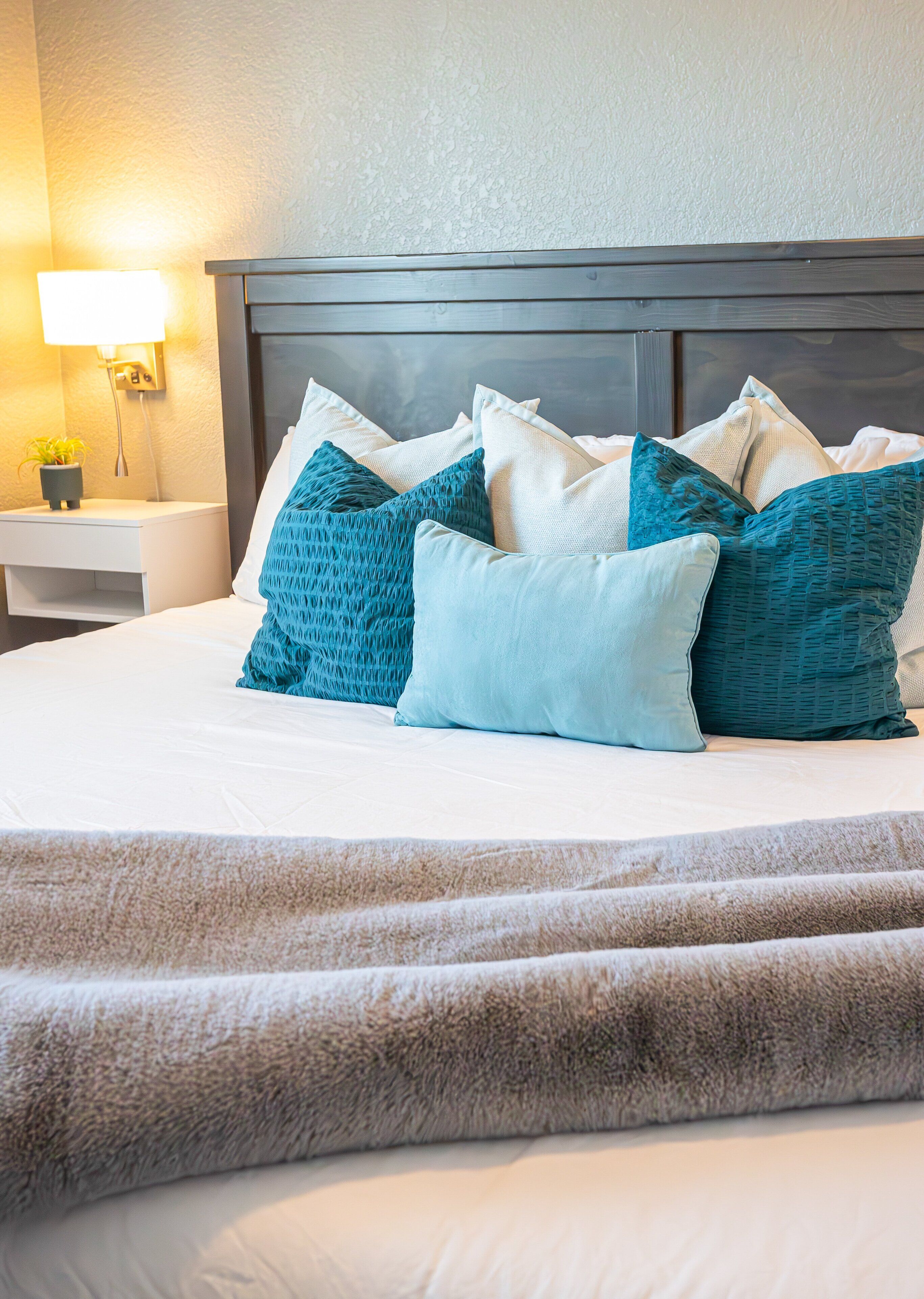 A neatly made bed with multiple pillows, a bedside table with a lamp, and a wall-mounted light fixture.