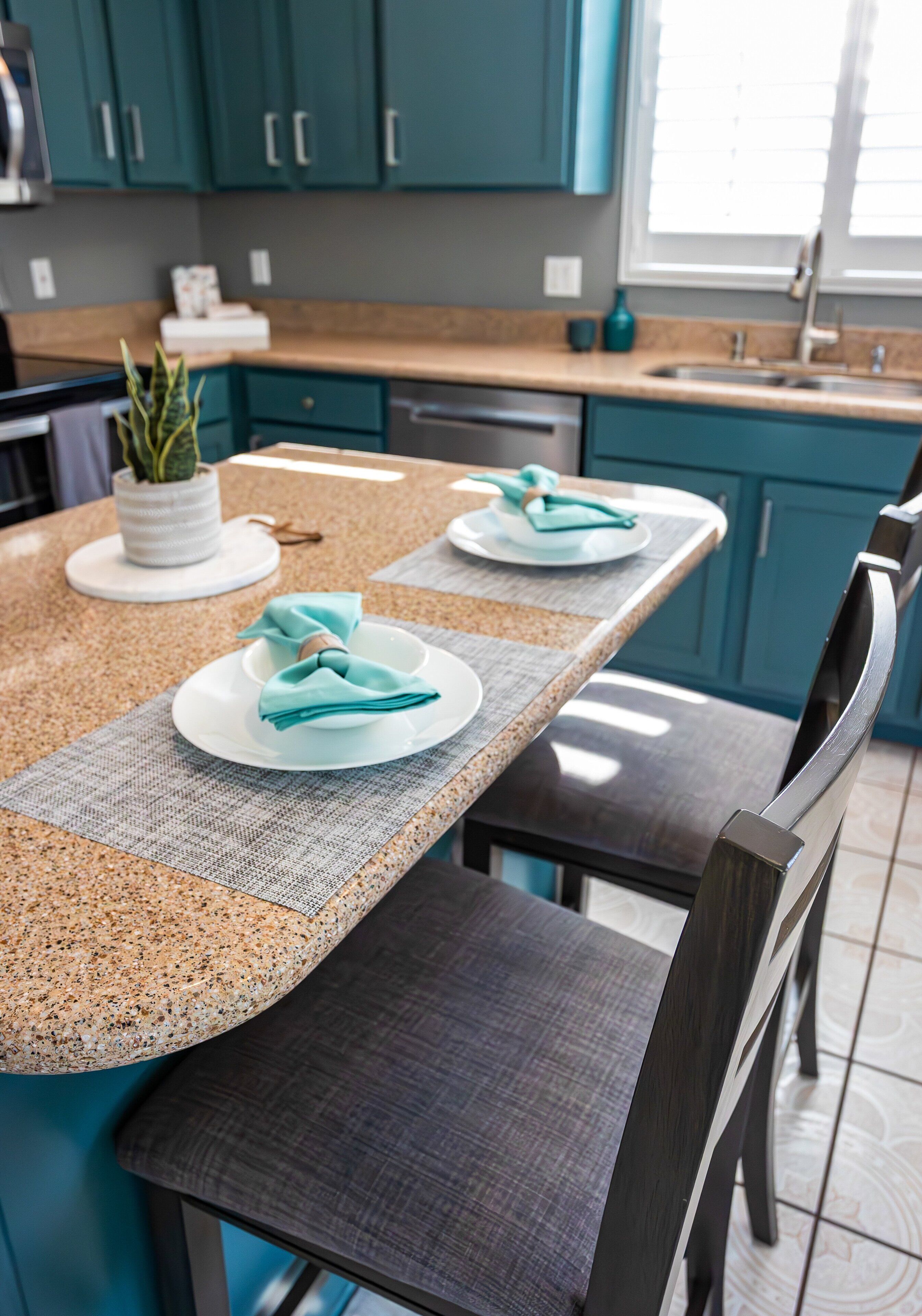 A kitchen with teal cabinets, granite countertops, and dark chairs.