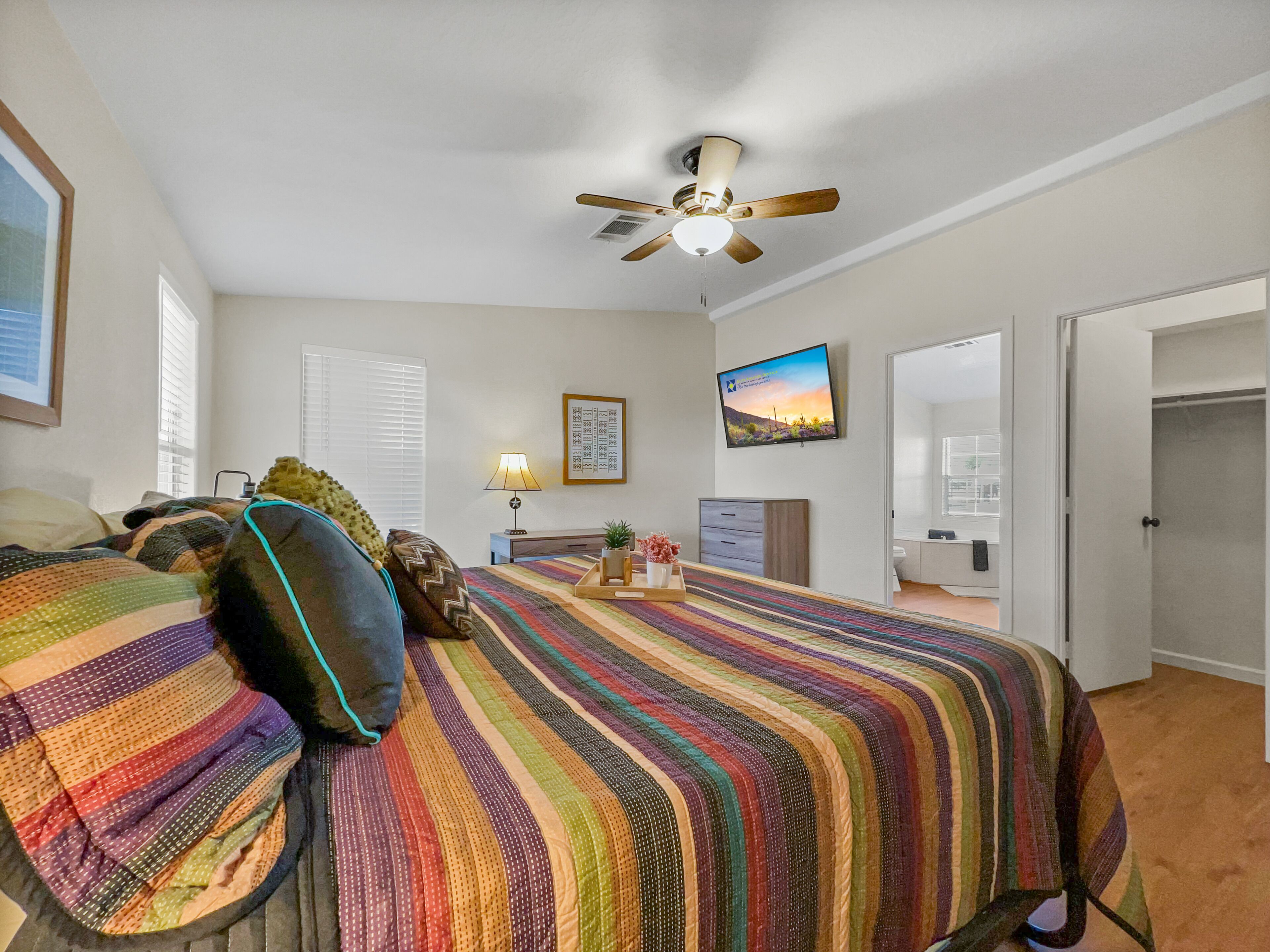 A bedroom with a large bed, a ceiling fan, a TV, and a door leading to another room.