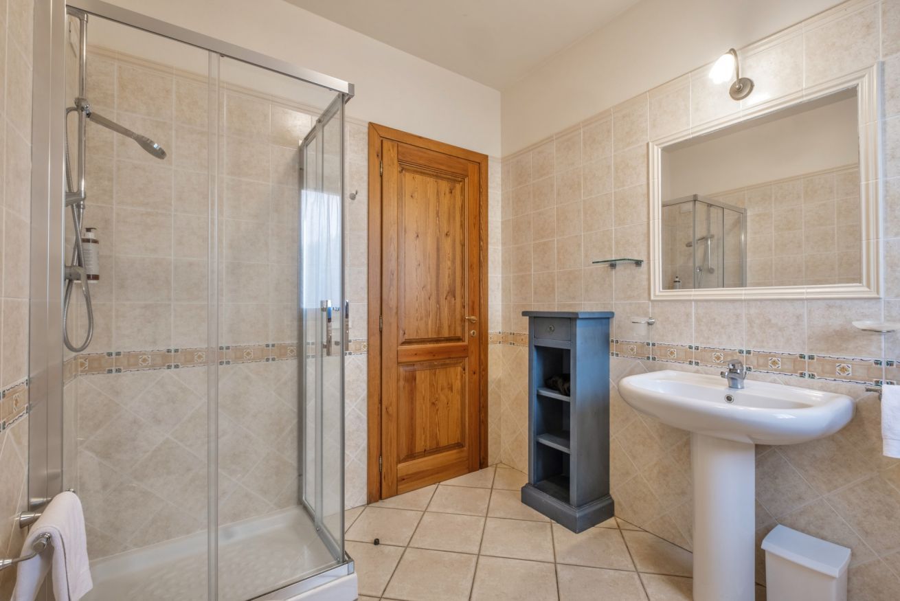 A bathroom with a glass shower enclosure, a wooden door, a pedestal sink, and a mirror above the sink.