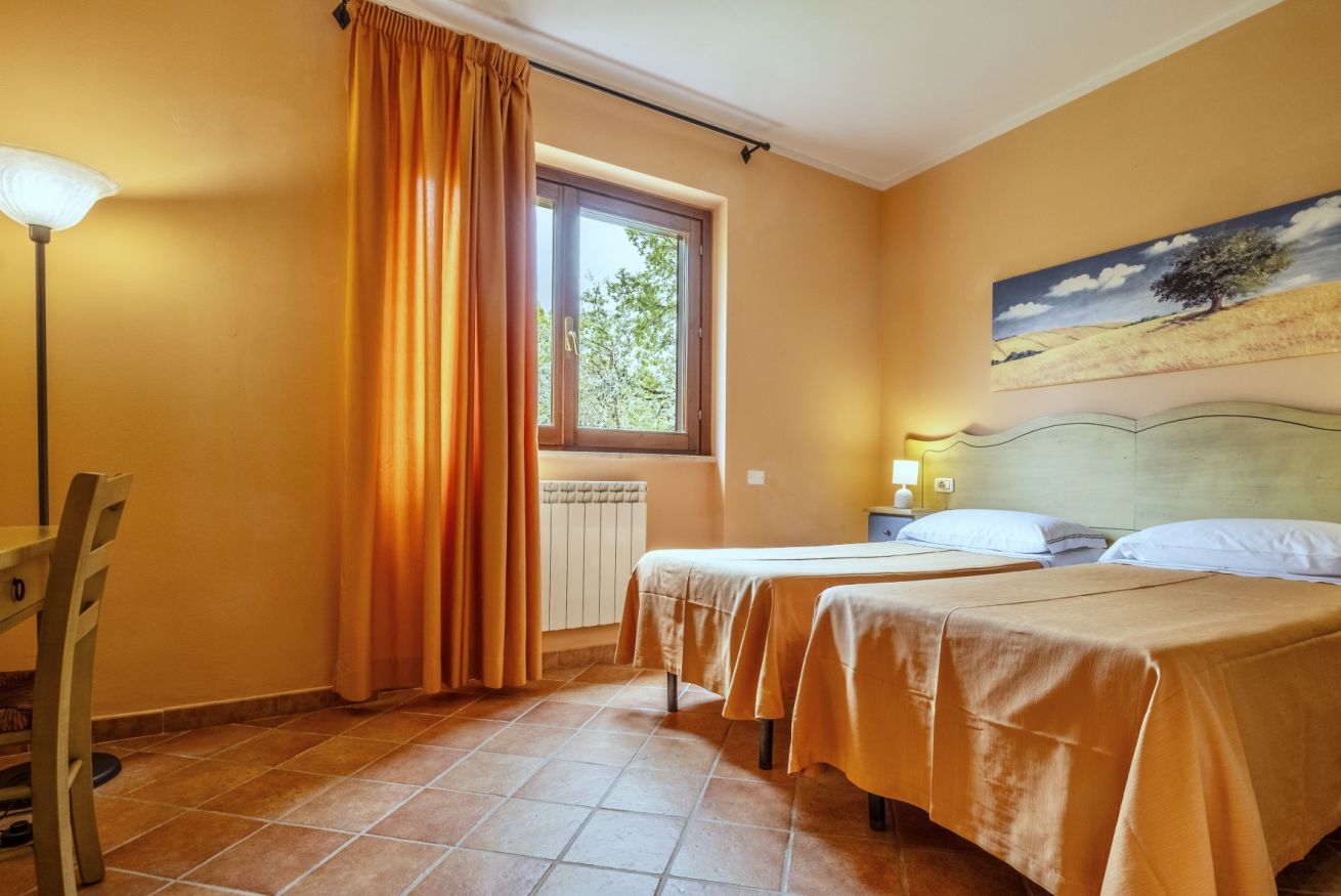 Une chambre d’hôtel avec deux lits, une fenêtre avec des rideaux, un radiateur, un tableau au mur et une lampe.