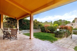 Een overdekt terras met een metselstenen vloer, een houten tafel en stoelen, en uitzicht op een verzorgde tuin met diverse planten en een stenen gebouw op de achtergrond.