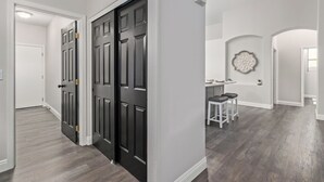 Un couloir avec des portes en bois foncé, un mur blanc orné d’une assiette décorative et un coin repas avec une table et des tabourets blancs.