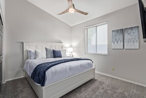 A bedroom with a white bed, a ceiling fan, a window with blinds, and two wall-mounted paintings. - Merino Mesa (Mesa)