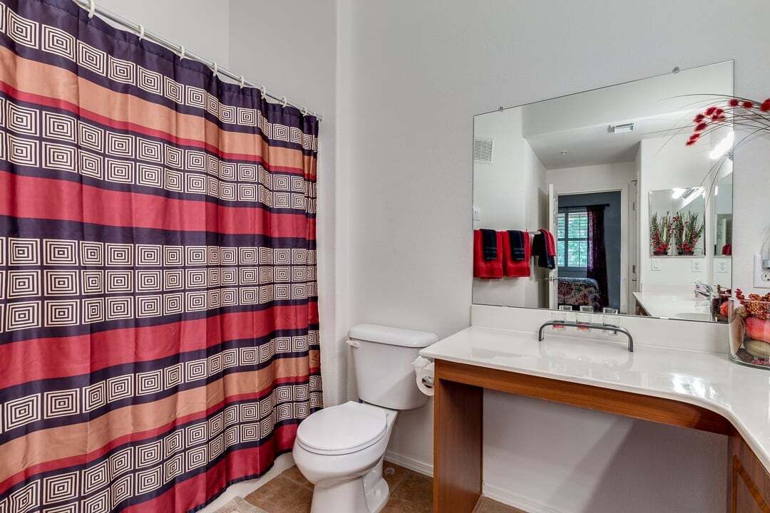 A bathroom with a white toilet, a white sink, and a mirror above it. There is a patterned shower curtain and a towel rack with red towels.
