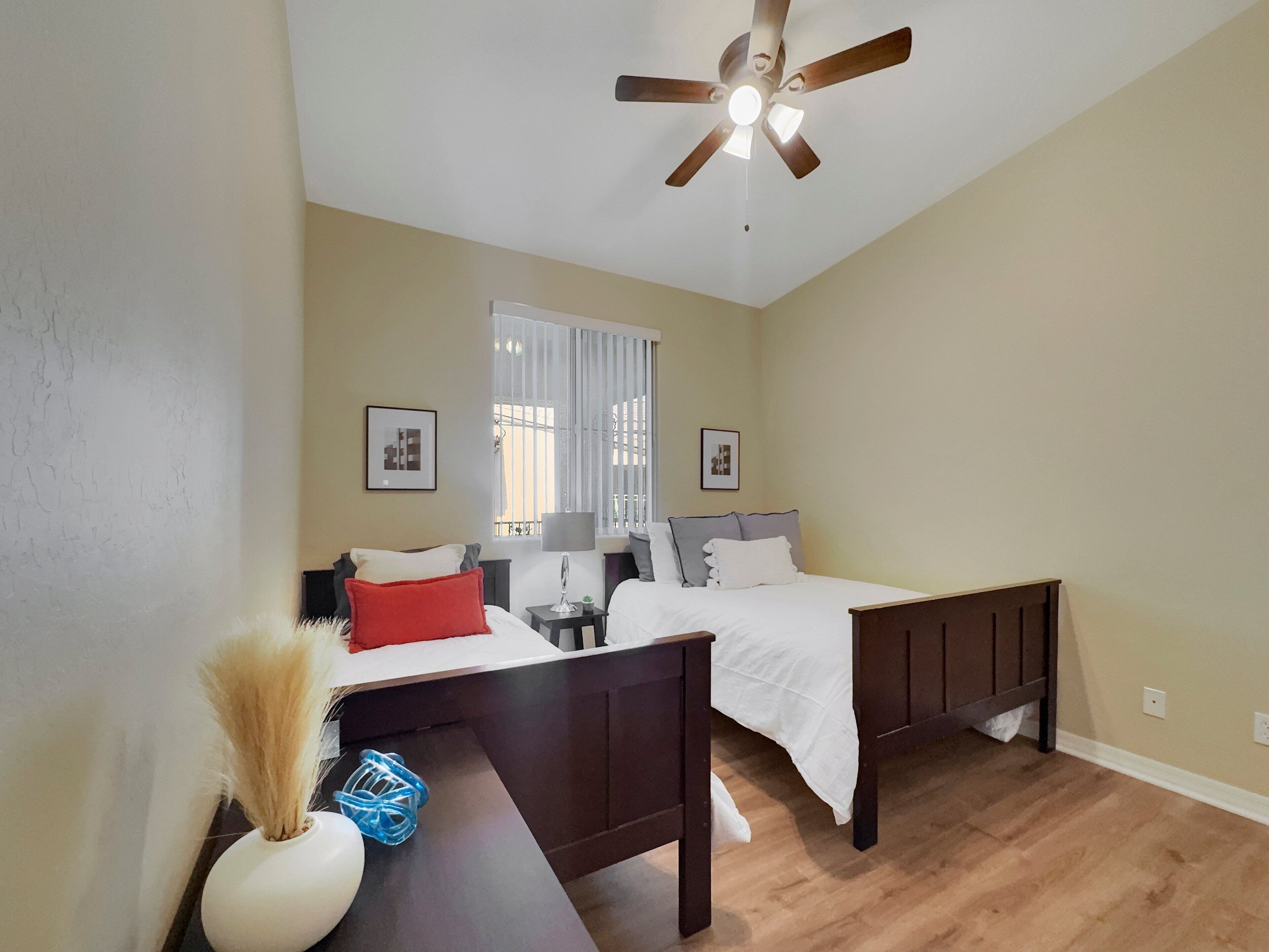 Une chambre avec deux lits, un ventilateur de plafond, une fenêtre avec des stores et un bureau avec une lampe et un vase.