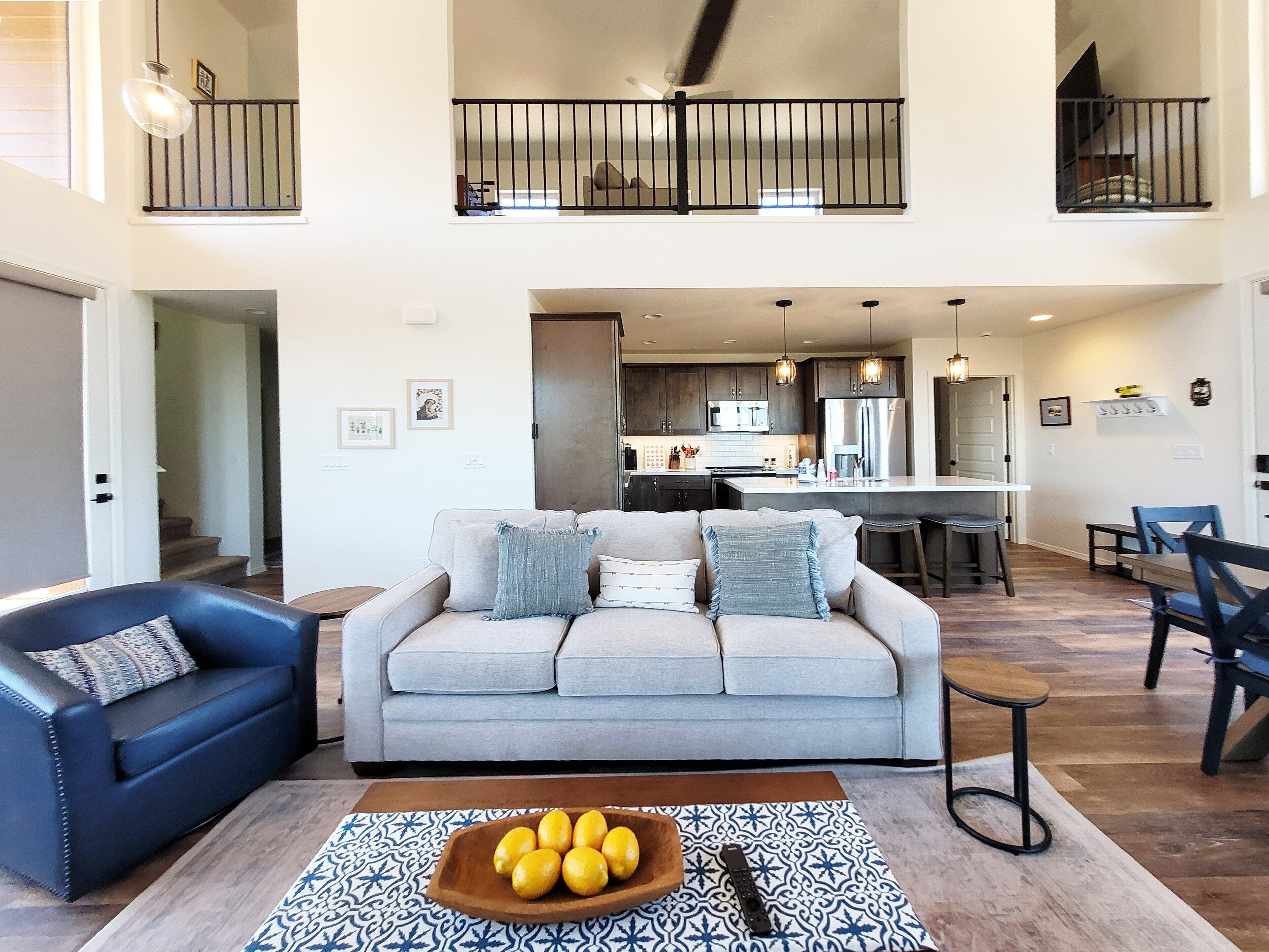 A spacious living room with a grey sofa, a blue armchair, and a wooden coffee table with lemons. A kitchen is visible in the background with modern appliances.