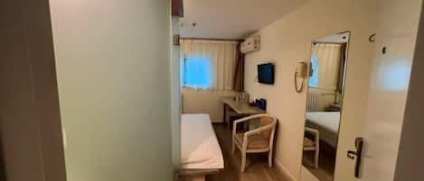 Una habitación de hotel pequeña con una cama individual, un escritorio, una silla, un espejo y una ventana con cortinas.