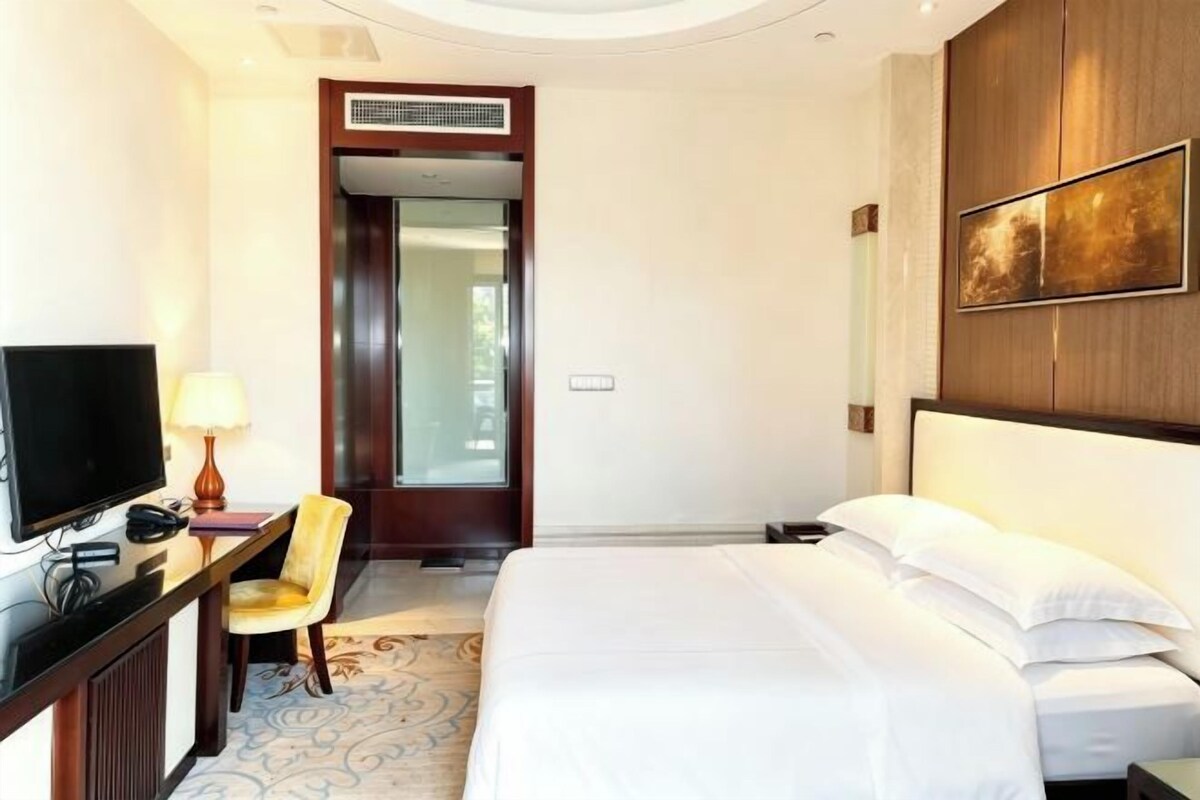 A hotel room with a bed, a desk with a television, a chair, and a door leading to another room.