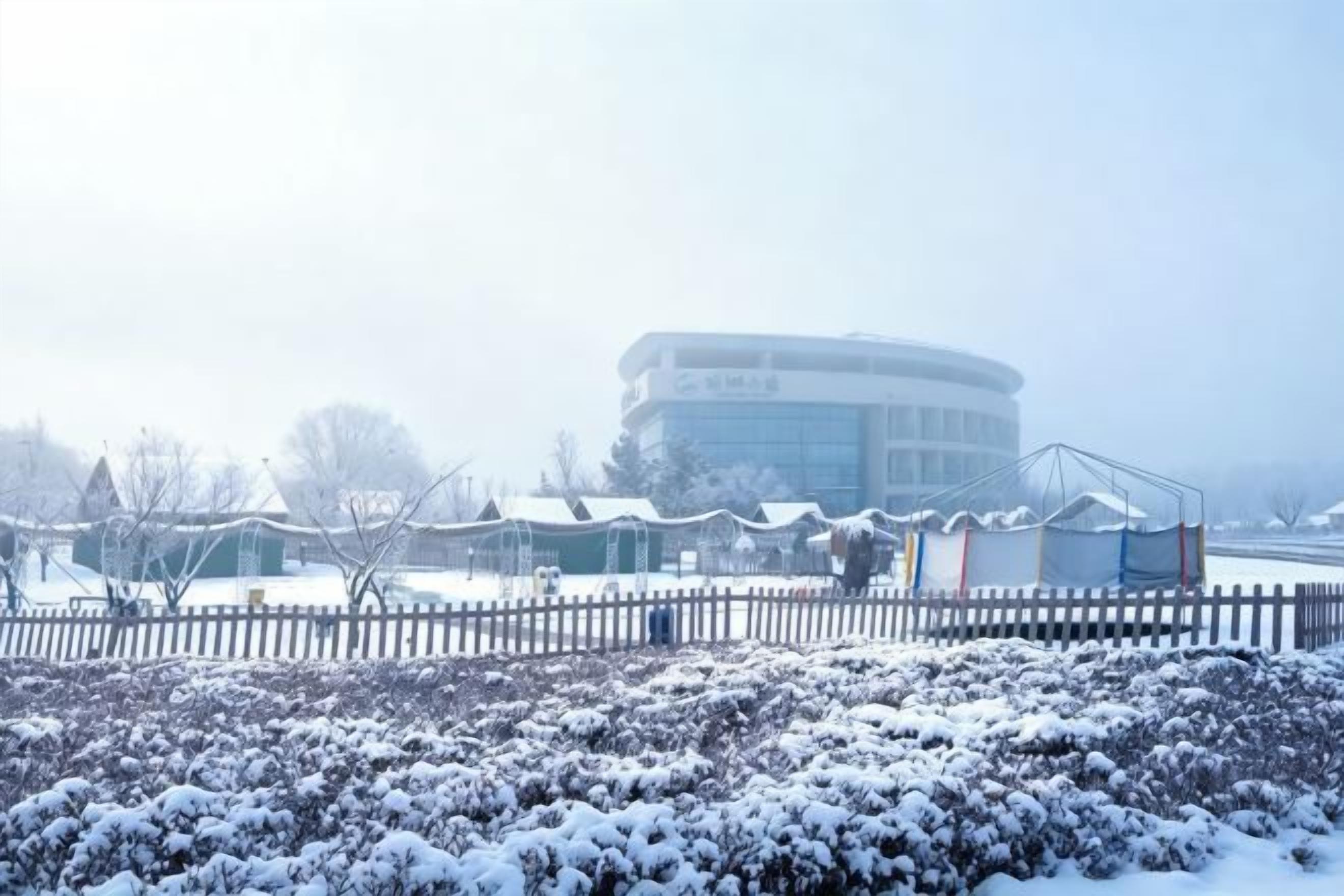 A building with a circular structure, a fenced area with snow-covered bushes, and a clear sky.