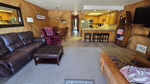 A cozy living room with a brown leather couch, a red chair, and a patterned rug. A kitchen with wooden cabinets and a bar area is visible in the background. - Lakeside Condo at Donner Lake - 2 Bdrm. 2 Bath - 1st floor - steps from the lake (Truckee)