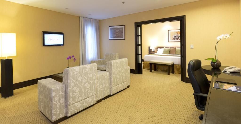A hotel room with a sofa, a desk with a phone, a TV, and a bed in the background.