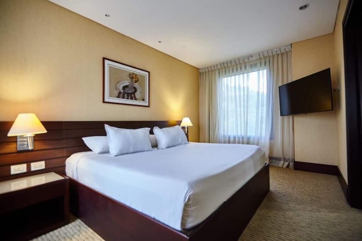 Une chambre d’hôtel avec un grand lit, deux tables de chevet avec des lampes, un téléviseur à écran plat fixé au mur et un tableau encadré au-dessus du lit.