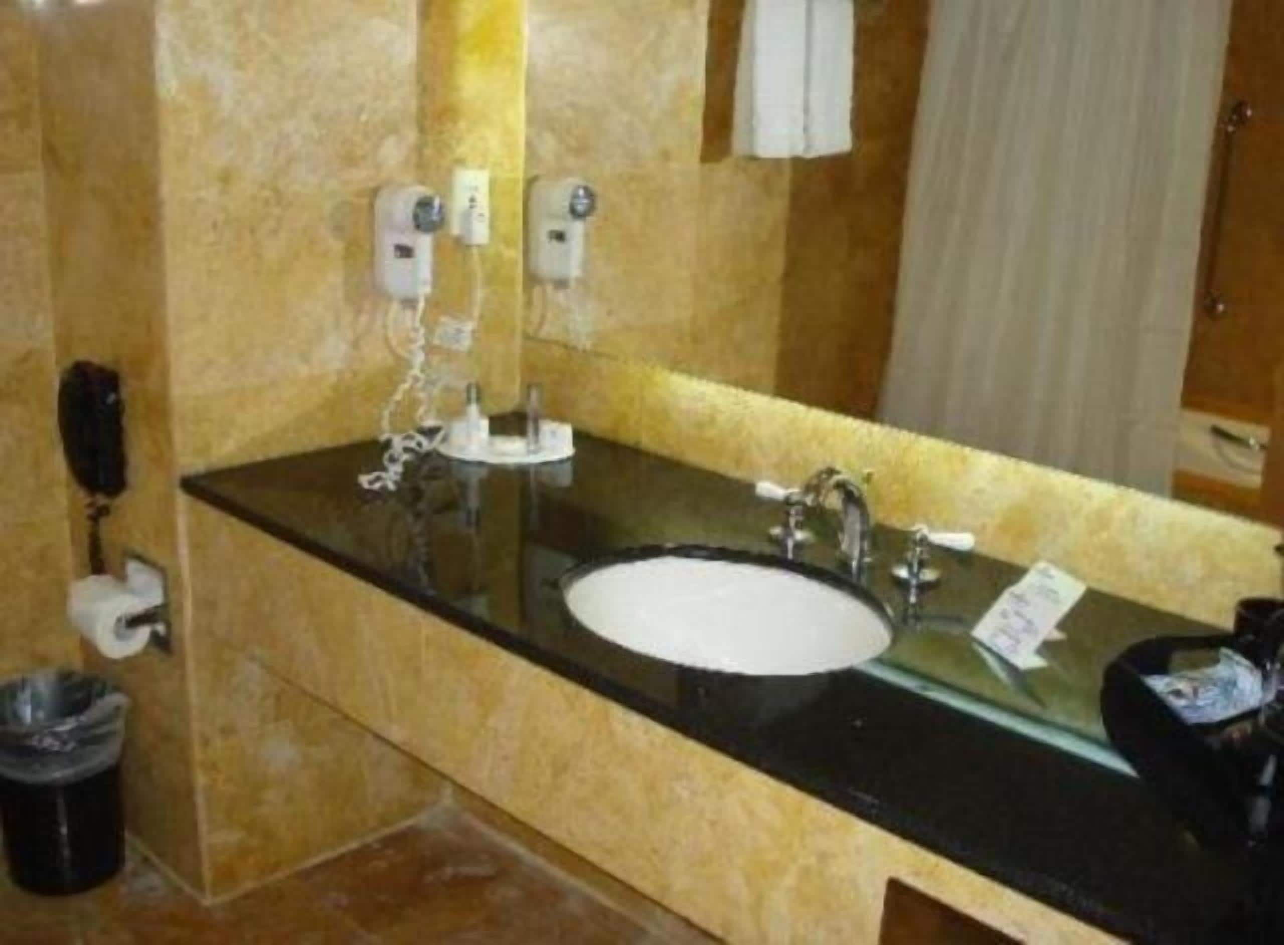 Une salle de bain avec un comptoir noir, un lavabo blanc et un miroir. On y trouve deux sèche-mains et une poubelle.