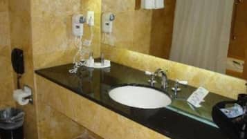 Une salle de bain avec un comptoir noir, un lavabo blanc et un miroir. On y trouve deux sèche-mains et une poubelle.