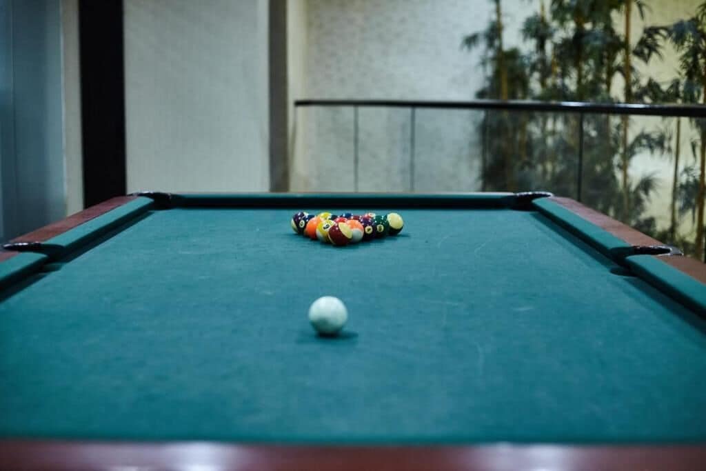 Una mesa de billar con una bola blanca y pelotas de colores, frente a un balcón con barandilla y vista a palmeras.